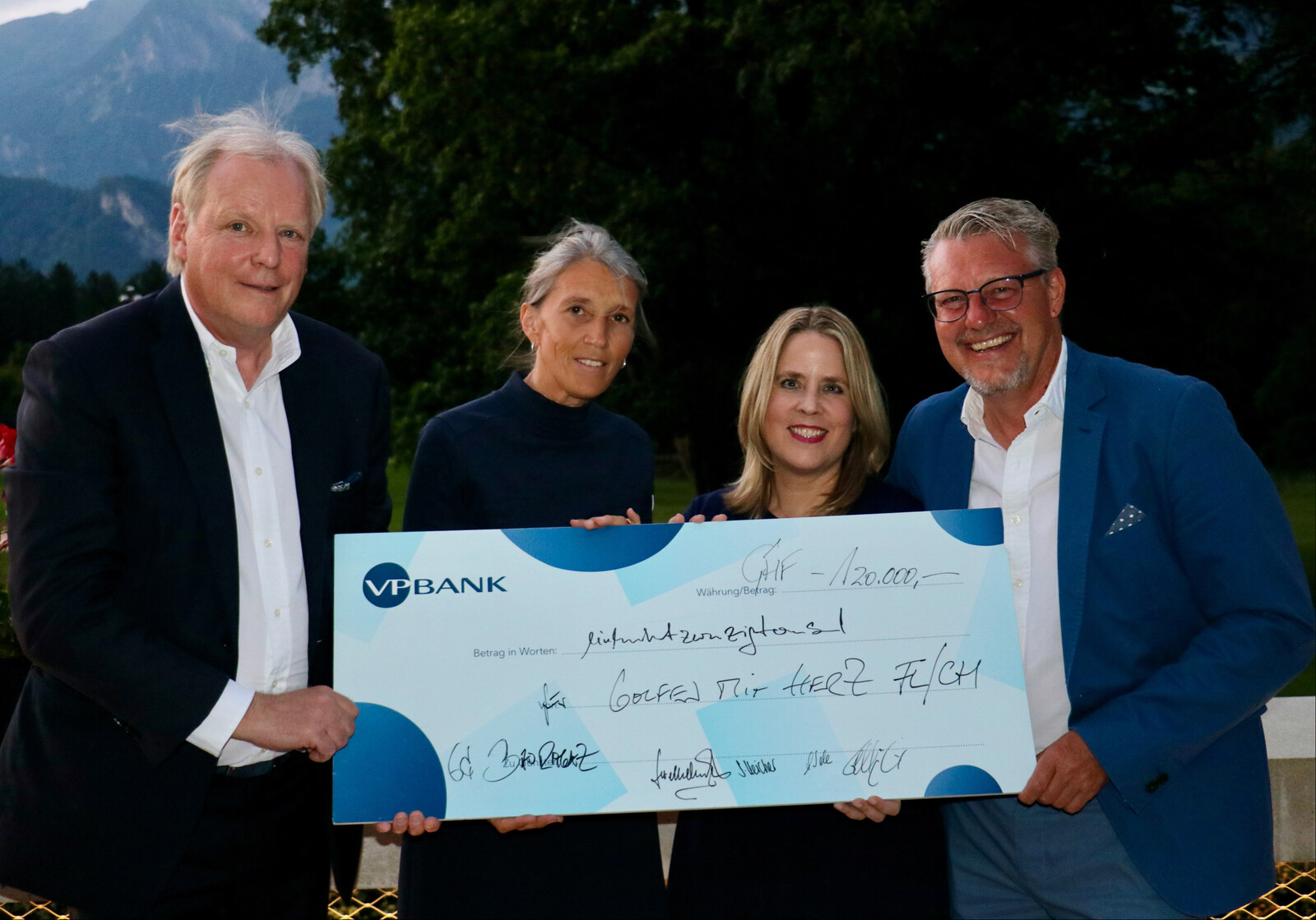 Vier lachende Menschen mit einem grossen VP Bank Check über 120'000 CHF im Freien, mit Bergen und Bäumen im Hintergrund. Die Stimmung ist feierlich.