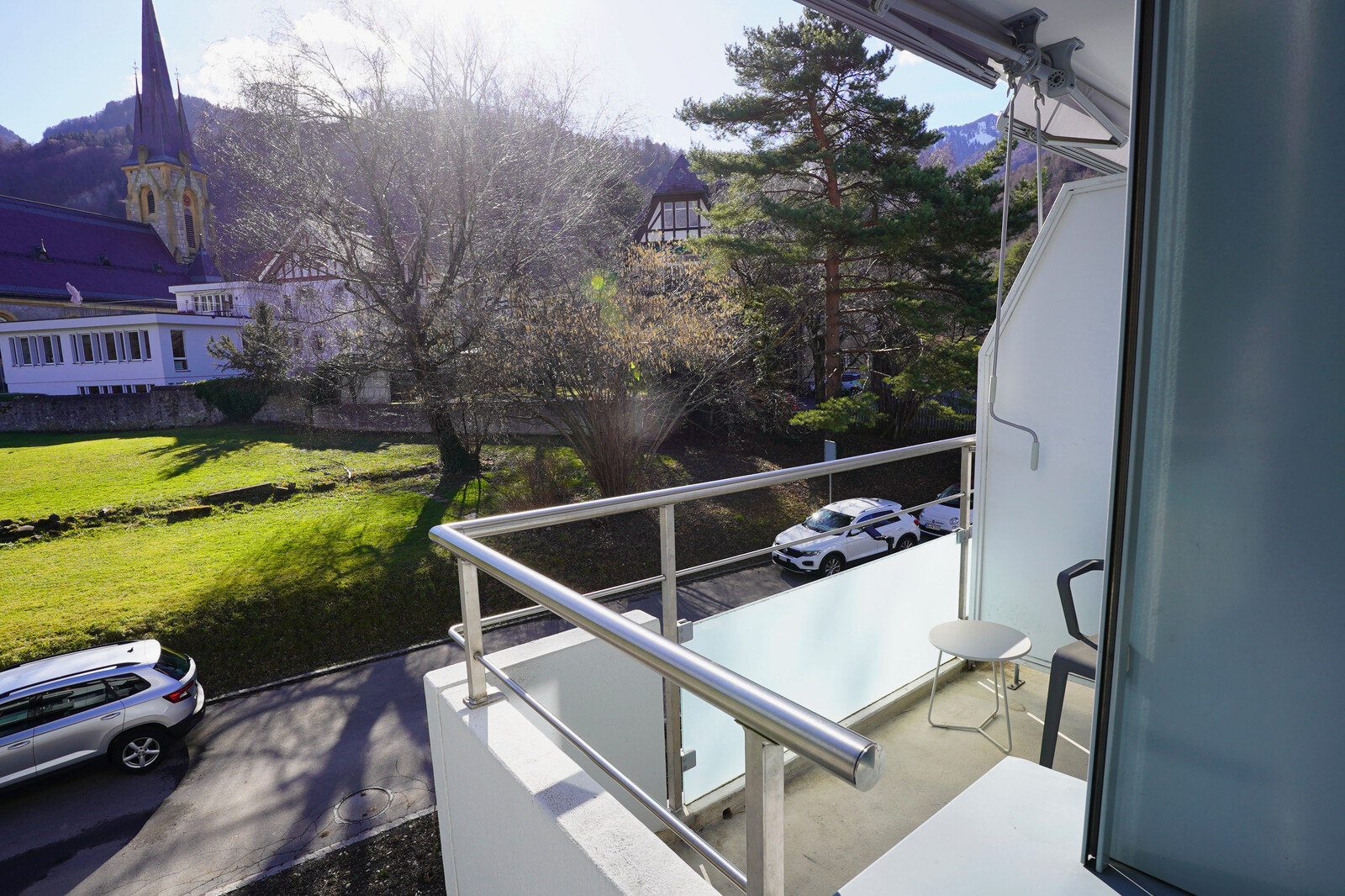 Sunlit balcony view with a chair and table. Overlooks a green yard, parked cars, tall pine trees, and a church with a mountain backdrop. Calm atmosphere.