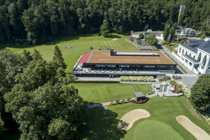 Luftaufnahme eines Golfplatzes inmitten einer üppigen Waldlandschaft. Im Vordergrund überragt ein moderne Clubhaus mit einer Terrasse mit rotem Dach die gepflegten Golfgrüns und Sandbunker und vermittelt eine friedliche, gehobene Atmosphäre.