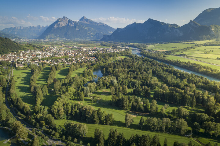 Luftaufnahme des 18-Loch Championship Course mit einem Fluss, der sich durch Felder und Bäume schlängelt. Eine kleine Stadt liegt am Fuße majestätischer Berggipfel unter einem klaren blauen Himmel.