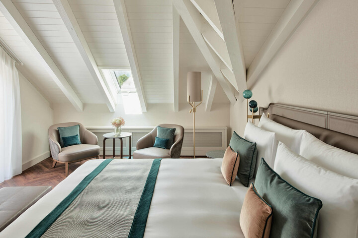 Gemütliches Schlafzimmer im Dachgeschoss mit schräger weißer Decke, einem ordentlich gemachten Bett mit tealfarbenen Akzenten, zwei Sesseln am Fenster.