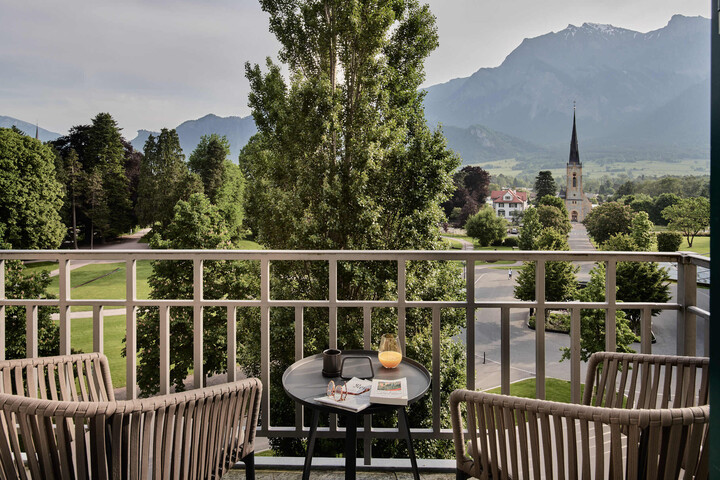 Balkon mit zwei Stühlen und einem Tisch mit Blick auf eine malerische Landschaft mit üppigen Bäumen, einer entfernten Kirchturmspitze und majestätischen Bergen unter einem bewölkten Himmel.