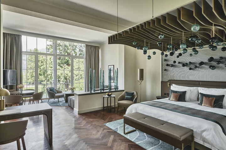 Luxuriöses Hotelzimmer vom Hof Ragaz mit einem grossen Bett, modernem Dekor und schwebenden Glaskugeln. Vom Boden bis zur Decke reichende Fenster geben den Blick auf das üppige Grün draußen frei und strahlen Ruhe aus.