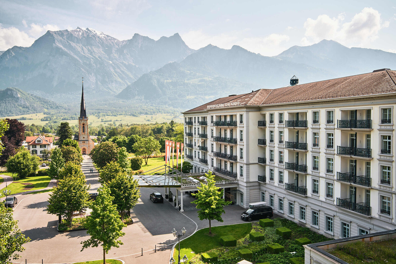 Das Grand Resort Bad Ragaz mit üppigen grünen Gärten und einer hohen Kirchturmspitze im Vordergrund. Majestätische Berge und klarer blauer Himmel im Hintergrund. Ruhiges Ambiente.