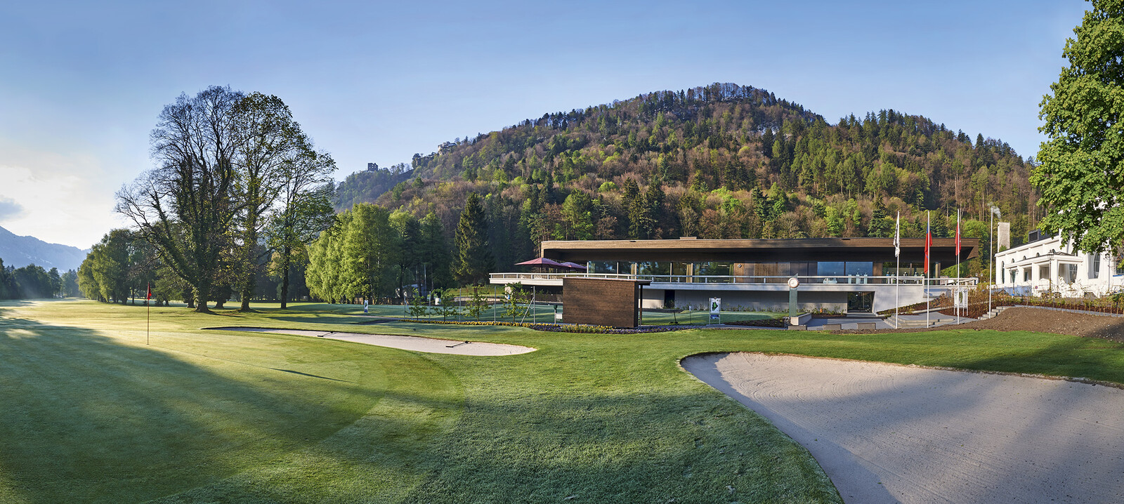 Der Goflplatz vom Golf Club Bad Ragaz inmitten eines bewaldeten Hügels. Das modern gestaltete Clubhaus ist umgeben von üppigem Grün unter einem klaren blauen Himmel.
