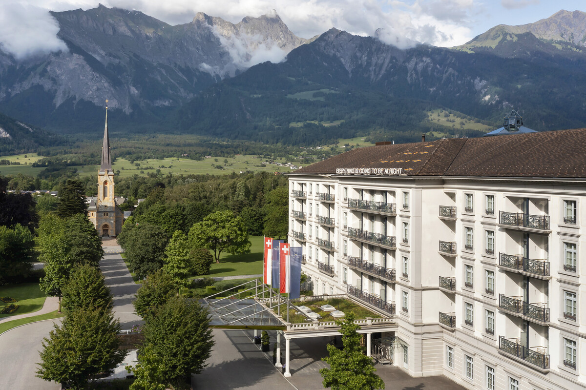 Seitenaufnahme des Grand Resort Bad Ragaz mit gestreiften Markisen und Balkonen vor einer Kulisse aus üppigem Grün und fernen Bergen unter einem teilweise bewölkten Himmel, der Ruhe ausstrahlt.