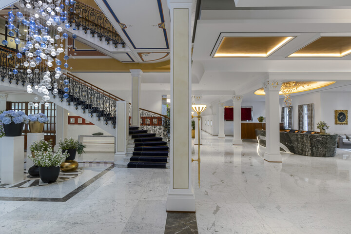 Luxuriöse Hotellobby des Grand Hotel Quellenhof mit grosser Treppe, kunstvollem Kronleuchter und Marmorböden. Elegante Sitzgelegenheiten und Dekoration sorgen für eine anspruchsvolle Atmosphäre.