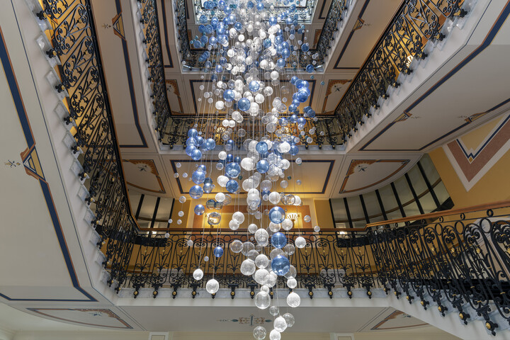 Die elegante, mehrstöckige Treppe des Grand Hotel Quellenhof mit verschnörkelten schwarzen Geländern wird von einem kaskadenförmigen Kronleuchter aus blauen und klaren Kugeln in Szene gesetzt, der ein anspruchsvolles Ambiente schafft.