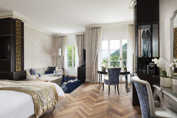 Geräumiges, elegantes Hotelzimmer mit einem gemütlichen Bett, gemustertem Teppich, Sofa, Schreibtisch und Stuhl. Grosse Fenster bieten einen malerischen Blick auf die Berge. Warme, einladende Atmosphäre.