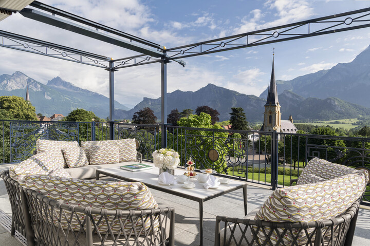 Die moderne Terrasse der King Suite mit gemütlichen Sitzgelegenheiten und einem Tisch für den Tee bietet einen Blick auf ein üppiges Tal und die fernen Berge. Ein Kirchturm verleiht dem Ganzen einen charmanten Touch.