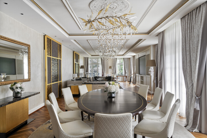 Elegantes Esszimmer mit einem runden Tisch, umgeben von weissen Stühlen, unter einem kunstvollen Kronleuchter mit goldenen Blättern. Große Fenster mit grauen Vorhängen, sanftes Tageslicht.