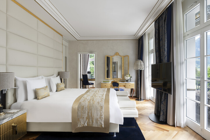 King Suite mit Kingsize-Bett und goldenen Akzenten. Große Fenster lassen Tageslicht herein. Ein Schreibtisch mit Spiegel und Fernseher sorgen für Eleganz und Komfort.