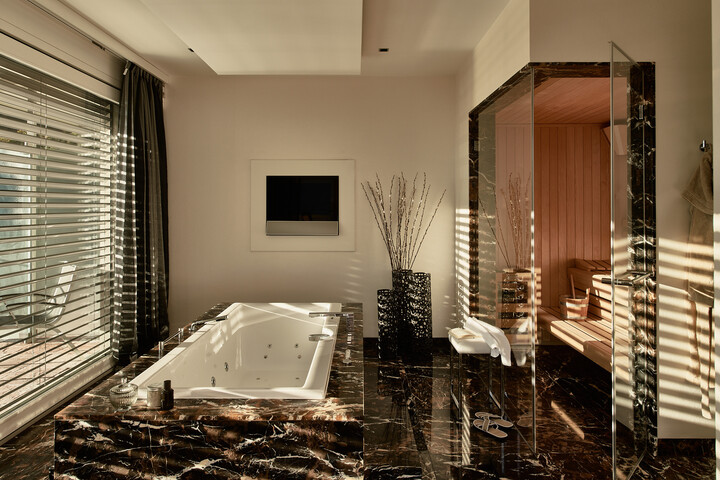 Luxuriöses Badezimmer der Penthouse Suite mit Marmor-Whirlpool, verglaster Sauna und großem Fenster mit Jalousien. Die warme Beleuchtung schafft eine ruhige, Spa-ähnliche Atmosphäre.