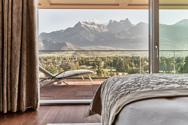 Schlafzimmer mit Bergblick. Im Vordergrund steht das Bett, bedeckt mit einer gemusterten beigen Decke. Auf dem Balkon steht ein Liegestuhl mit Blick auf grüne Felder und majestätische schneebedeckte Gipfel.