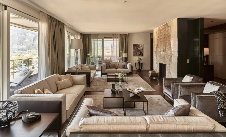 Luxuriöses Wohnzimmer der Penthouse Suite mit beigen Sofas, grossen Fenstern und Blick auf die Berge. Elegante Einrichtung, sanfte Beleuchtung, weiche Teppiche, die eine warme, einladende Atmosphäre vermitteln.