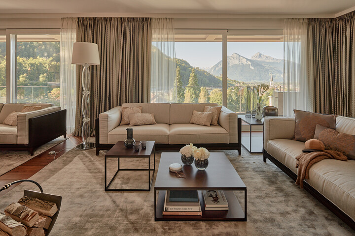Elegantes Wohnzimmer der Penthouse Suite mit beigen Sofas und Kissen in warmen Farbtönen. Große Fenster bieten einen Blick auf die Berge. Auf den Couchtischen liegen Bücher und Blumen, die eine ruhige, gemütliche Atmosphäre schaffen.