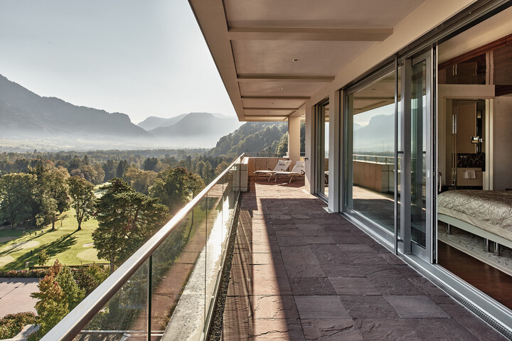 Geräumiger Balkon der Penthouse Suite mit Glasgeländer und Blick auf ein ruhiges Tal und die Berge. Modernes Interieur durch große Glastüren sichtbar. Friedliche, sonnige Atmosphäre.