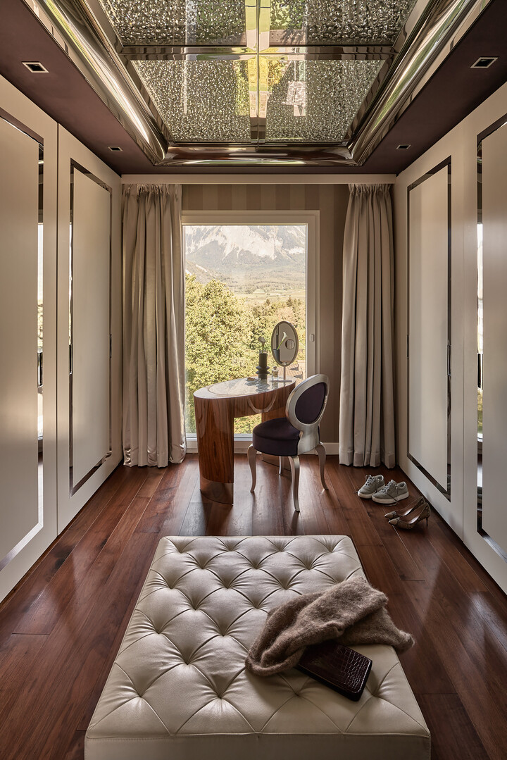 Luxuriöses Ankleidezimmer mit gepolstertem Ottoman, Holzkommode und elegantem Stuhl. Grosses Fenster mit Bergblick, Schuhen und einer kuscheligen Decke.