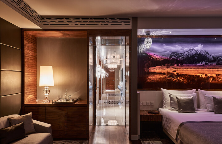 Modernes Zimmer der Presidential Suite mit warmer Beleuchtung, gemütlichem Bett, Bergwandbild, eleganter Lampe und glänzendem Flur, in dem sich Kronleuchter spiegeln.