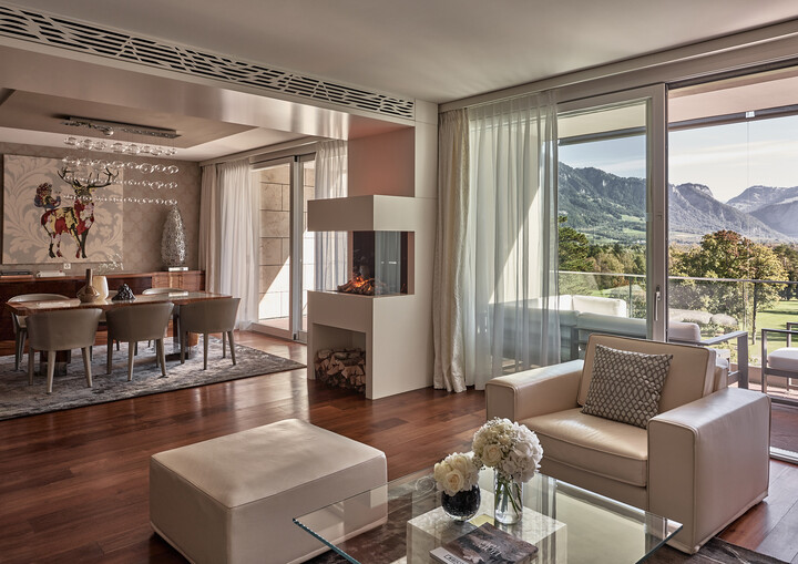 Luxuriöses Wohnzimmer mit Blick auf die Berge. Mit großen Fenstern, gemütlichem Kamin, stilvollen beigen Sitzmöbeln, elegantem Essbereich und Hirschkunstwerken.