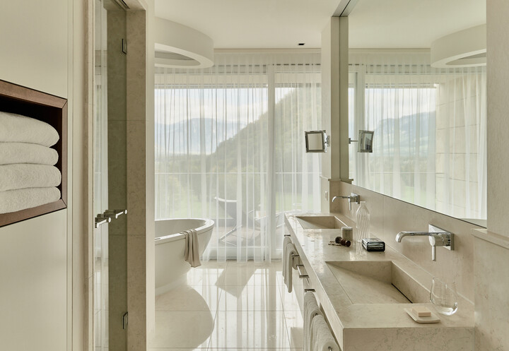 Modernes Badezimmer der Spa Suite mit grossem Spiegel, zwei Waschbecken und Steinwaschtisch. Eine freistehende Badewanne steht neben Fenstern mit transparenten Vorhängen und bietet einen Blick auf die Berge.