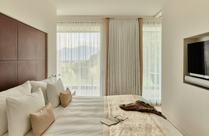 Helles, modernes Schlafzimmer der Spa Suite mit grossen Fenstern, transparenten Vorhängen und Blick auf die Berge. Das Bett mit weichen Kissen, einem Buch und einer Brille schafft eine gemütliche, ruhige Atmosphäre.