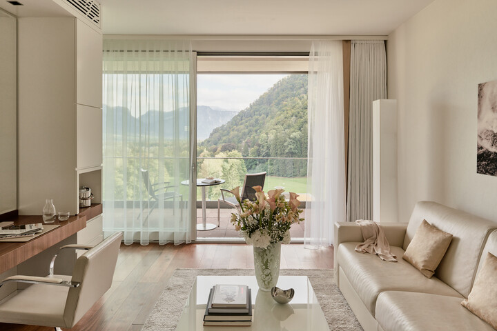 Modernes Wohnzimmer der Spa Suite mit beigem Sofa, Holzakzenten und einer Blumenvase auf dem Couchtisch. Das große Fenster bietet einen herrlichen Blick auf die Berge und schafft eine ruhige, gemütliche Atmosphäre.