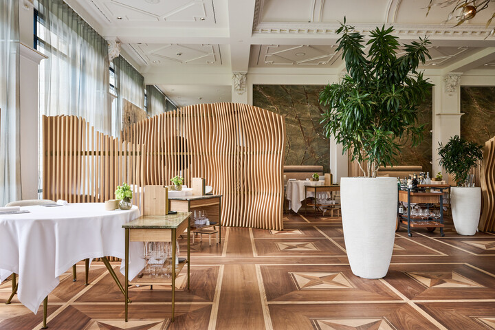 Elegantes Restaurantinterieur des Restaurant Sven Wassmer Memories mit grossen Grünpflanzen in weißen Pflanzkübeln, Trennwänden aus Holz und Tischen mit weißen Tischdecken. Die Atmosphäre ist ruhig und gediegen.