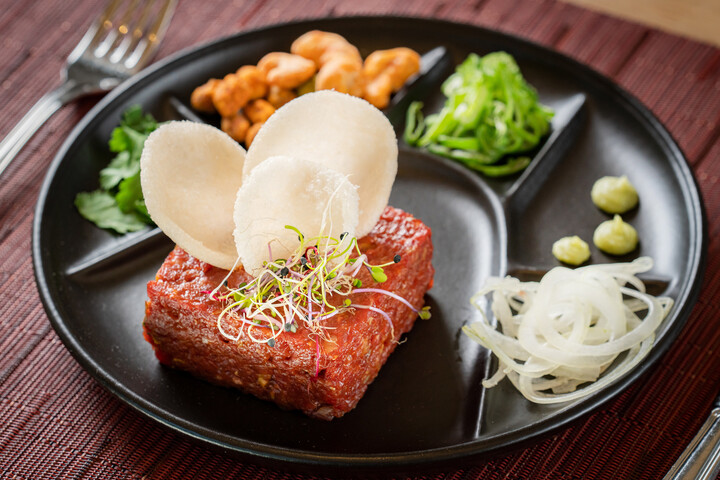 Ein schwarzer Teller mit einem Tartar-Block, garniert mit Sprossen und Garnelencrackern. Darum herum liegen Cashewnüsse, geschnittenes Gemüse, Zwiebeln und Wasabi. Elegant und appetitlich.