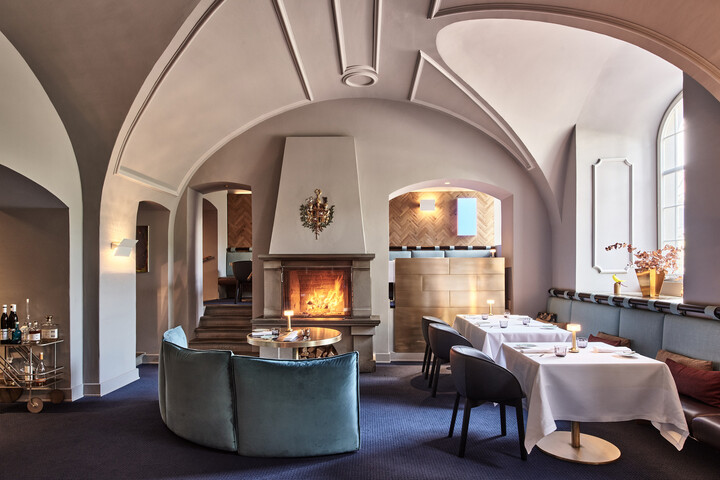 Der Innenbereich des Restaurant Igniv wird gezeigt. In der Mitte brennt Feuer in einem Kamin. Davor steht ein Sofa. Auf der rechten Seite stehen Tische und Stühle. Das Restaurant befindet sich im Grand Resort Bad Ragaz, einem Sternehotel in der Schweiz.