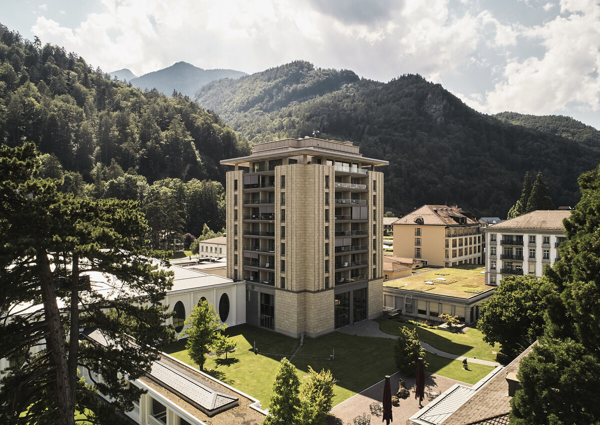 Der moderne Spa Tower in Bad Ragaz mit Balkonen steht inmitten üppiger Vegetation und ist von bewaldeten Bergen umgeben, während der Himmel teilweise bewölkt ist.