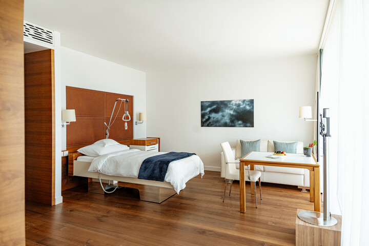 Modernes Hotelzimmer des Grand Resort Bad Ragaz in einem ruhigen, minimalistischen Design. Ausgestattet mit einem Bett, einem Sofa, einem Fernseher und einem Tisch mit rosa Blumen. Das bodentiefe Fenster führt auf einen Balkon mit Panoramablick.