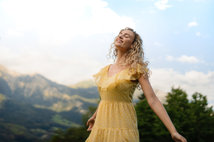 Eine Frau in einem gelben Kleid hebt vor dem Hintergrund grüner Hügel und blauem Himmel freudig ihre Arme. Ihr Gesichtsausdruck ist gelassen, sie umarmt die Natur.
