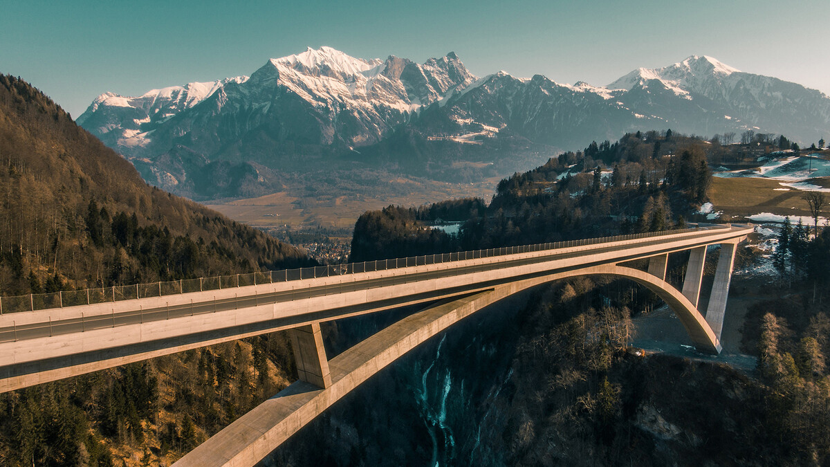 The Tamina Bridge can be seen in the middle. The sun is setting. Mountains and forests surround the bridge. Nearby is the Superior Hotel Grand Resort Bad Ragaz, a star hotel in Switzerland.