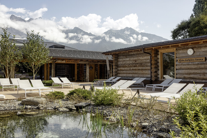 Malerisches Saunadorf in den Bergen mit Holzsaunen und weißen Liegestühlen an einem Teich. Die hohen Berge im Hintergrund sind von einer sanften Wolkendecke bedeckt, was eine ruhige Atmosphäre schafft.