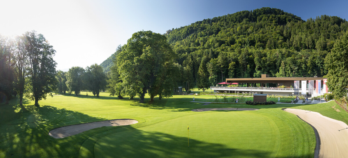Das Golfclubhaus ist in seiner gesamten Länge zu sehen. Die Sonne leuchtet seitwärts in die Kamera. Bäume und der Golfrasen befinden sich vor dem Golfclubhaus.
