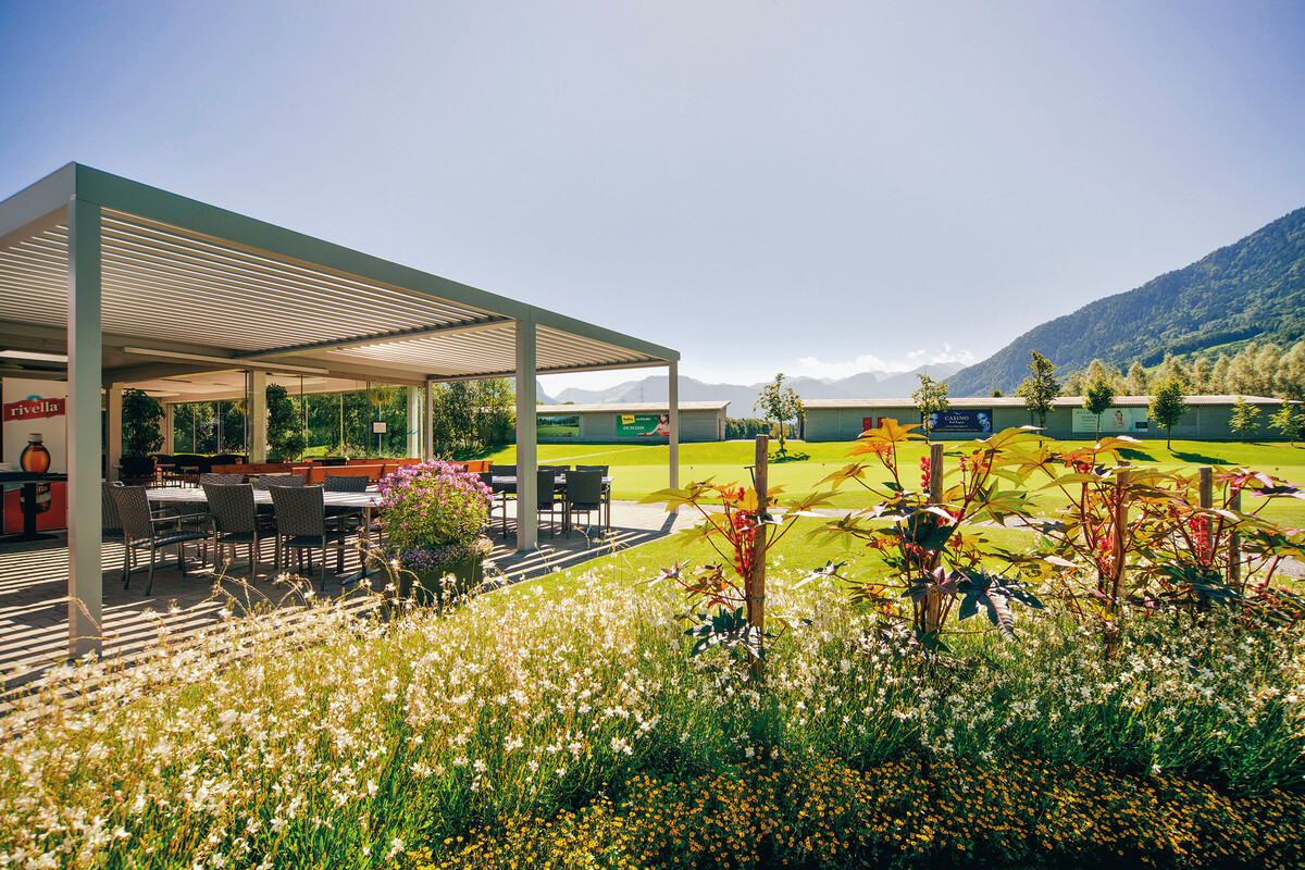 Geräumige Aussenterrasse vom Golf Bistro mit moderner Pergola, umgeben von blühenden Blumen und üppigem Grün. Die Berge im Hintergrund unter einem hellen, klaren Himmel.