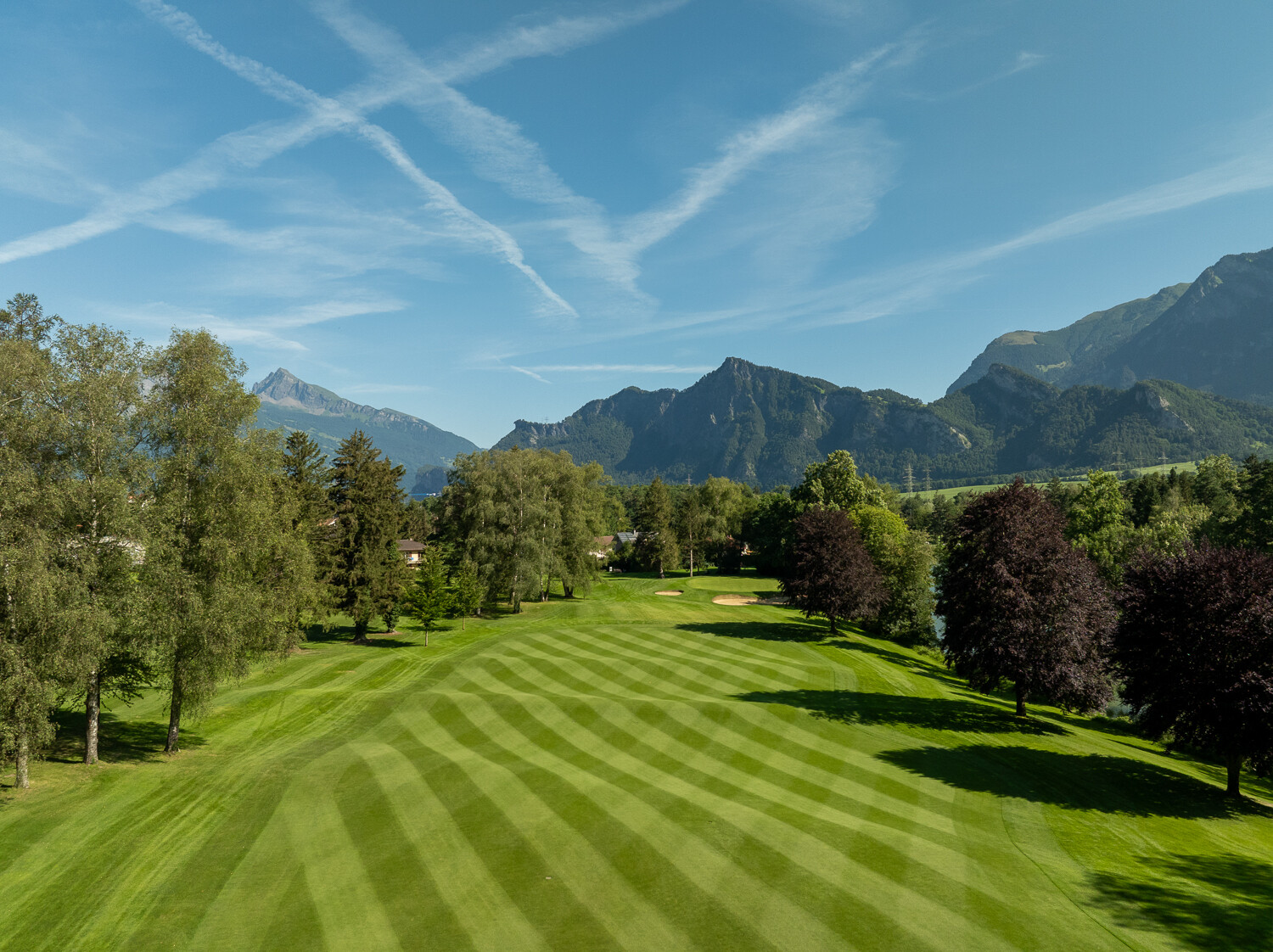 Der Golf Platz Bad Ragaz mit üppig grünen Fairways und Bäumen, unter einem klaren blauen Himmel mit Wolkenfetzen. Die Bergkette bietet eine majestätische Kulisse.
