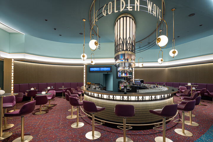 Eine Bar im Retro-Stil mit dem Schild „The Golden Wave“. Lila Stühle umgeben eine runde Bar mit eleganter Beleuchtung. Gemütliche, lebhafte Atmosphäre mit Art-déco-Details.