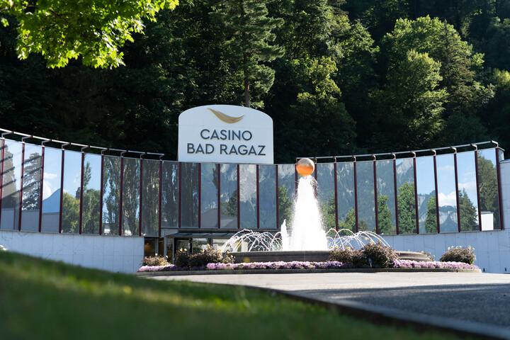 Das moderne Gebäude des Casino Bad Ragaz mit der Aufschrift „Casino Bad Ragaz” und verspiegelten Wänden, die Bäume reflektieren. Ein zentraler Springbrunnen und üppiges Grün schaffen eine einladende Atmosphäre.