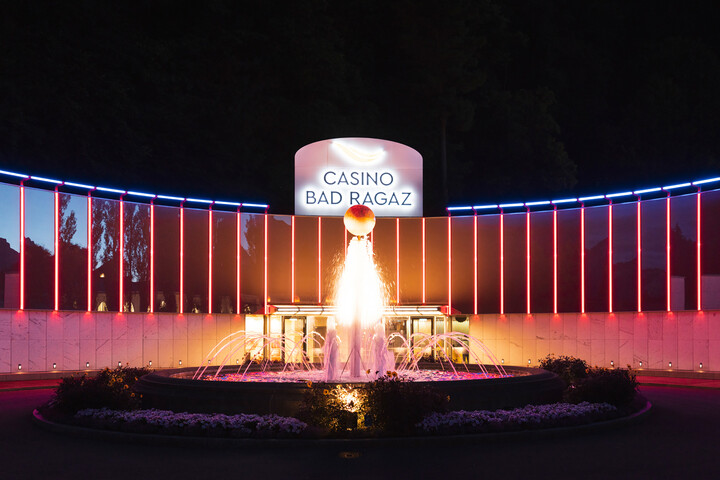 Nachtansicht des Casinos Bad Ragaz mit einem hell erleuchteten Brunnen davor. Neonrote und blaue Lichter betonen das moderne, lebendige Design des Gebäudes.