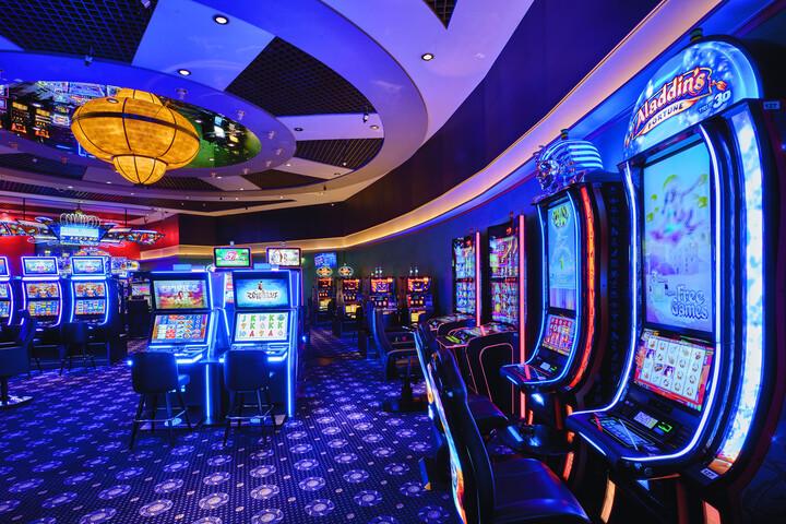 Das lebhafte Casino-Interieur des Casino Bad Ragaz mit Reihen hell erleuchteter Spielautomaten unter einer grossen, dekorativen Deckenlampe. Die Atmosphäre ist lebhaft und farbenfroh.