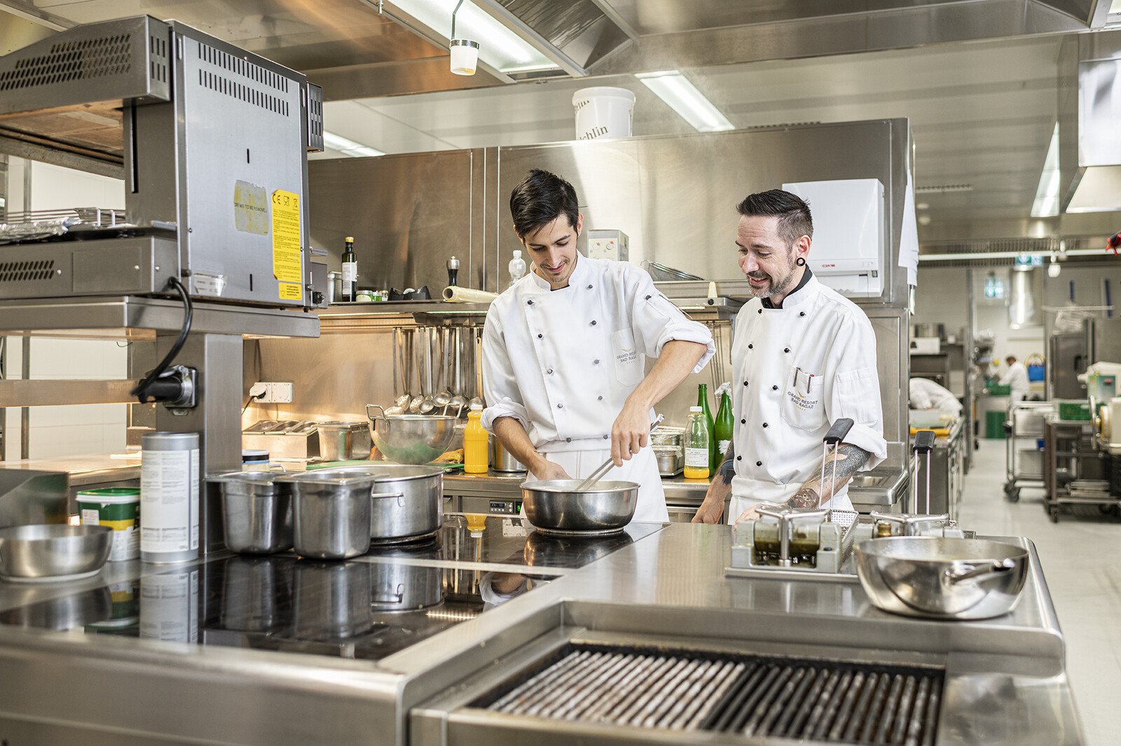 Vier Köche stützen sich, in ihrer Uniform, auf dem Küchentresen ab. In ihrem Hintergrund ist die Küche zu sehen. Die Küche gehört zu dem Hotel Grand Resort Bad Ragaz.