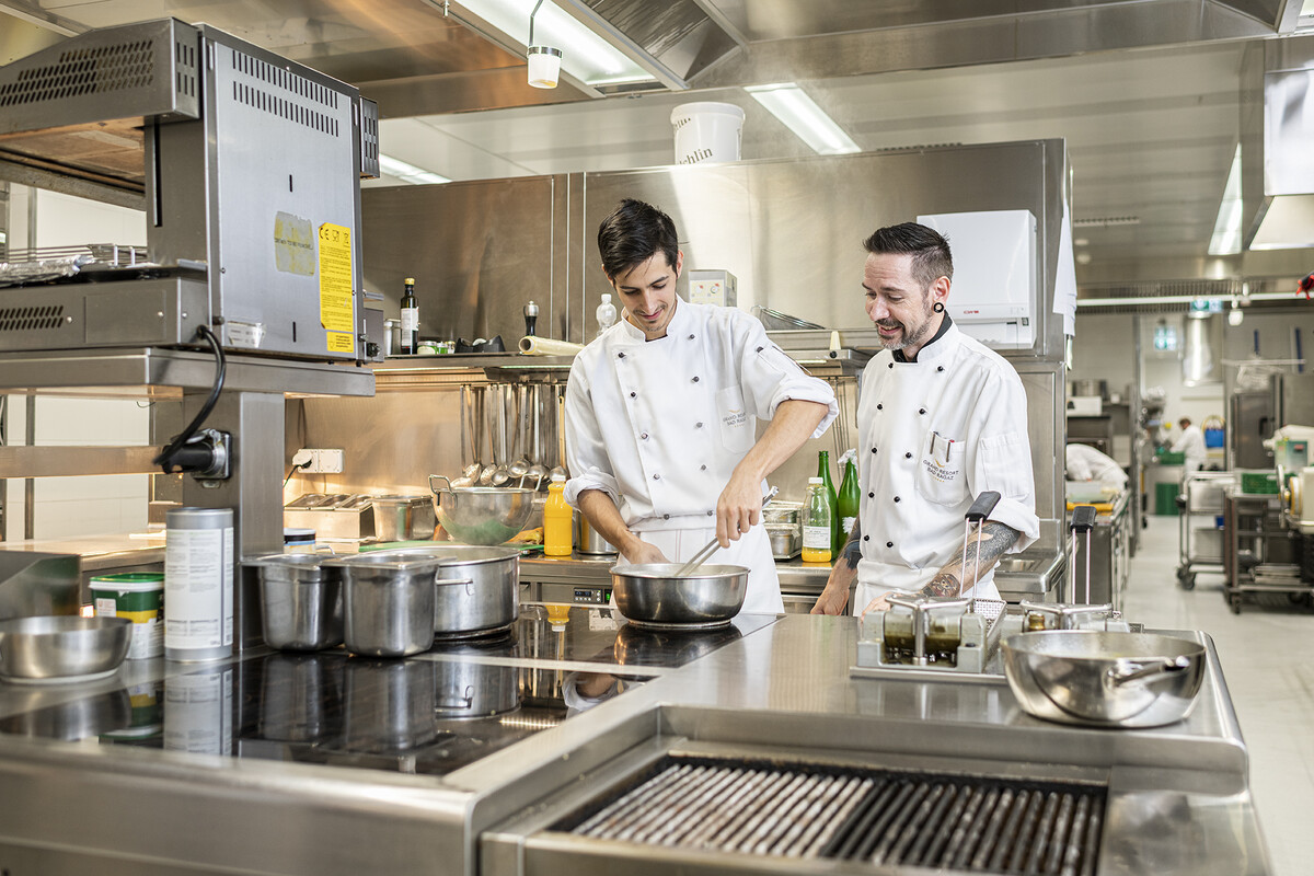 Four chefs, in their uniforms, lean on the kitchen counter. The kitchen can be seen in the background. The kitchen belongs to the Hotel Grand Resort Bad Ragaz.
