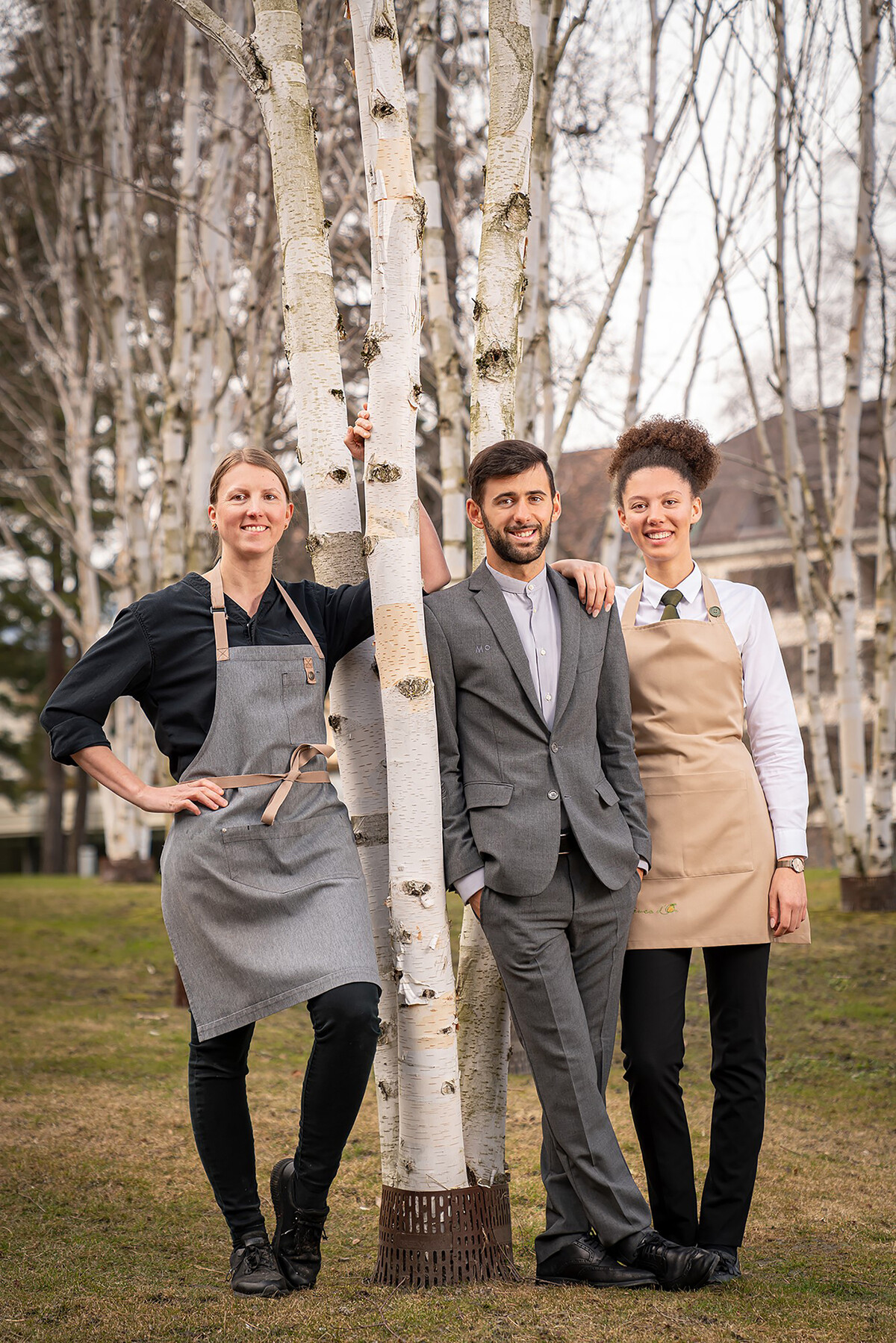 Drei Lernende aus unterschiedlichen Bereichen lehnen draussen an einem Baum. Hinter ihnen ist ein Teil des Hotel Grand Resort Bad Ragaz zu sehen, einem Sternehotel in der Schweiz.