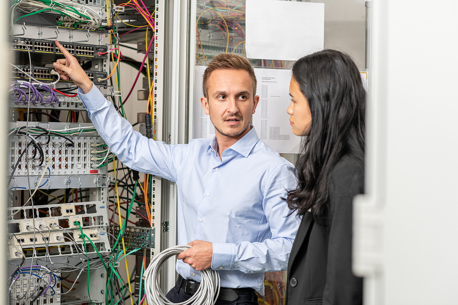 Der Ausbildner zeigt der Lernenden der Infrastruktur einige Kabel. Sie befinden sich vor einem Kasten mit vielen Kabeln. Sie sind im Hotel Grand Resort Bad Ragaz angestellt.