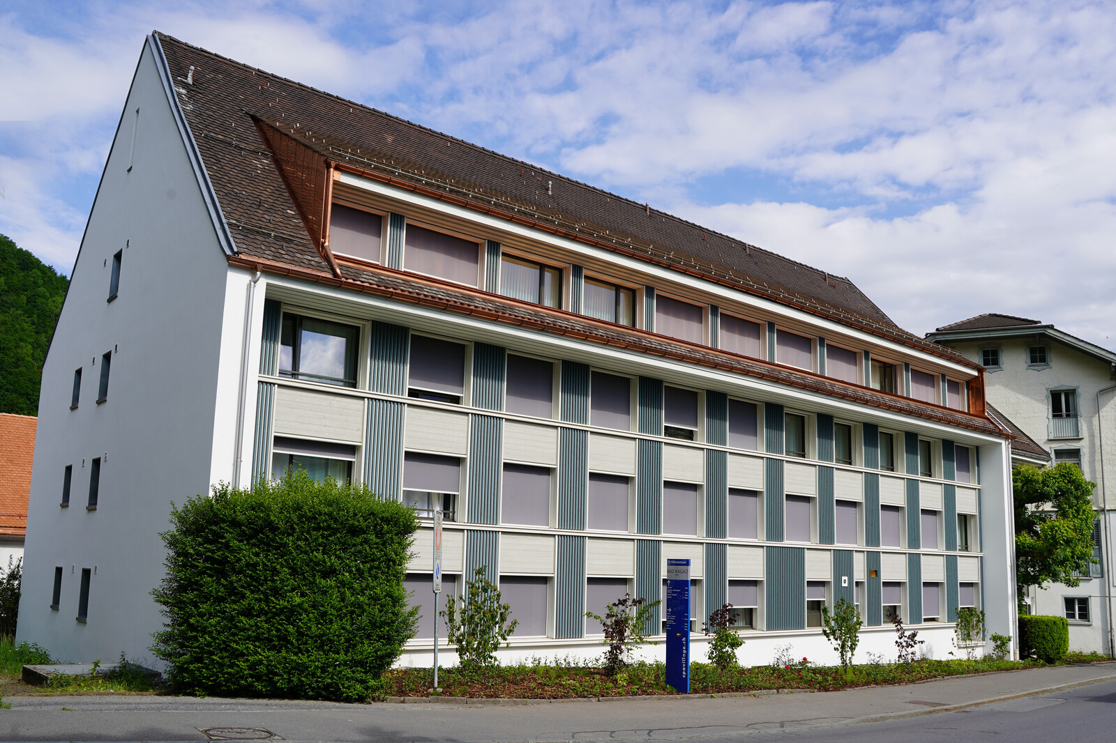 Modernes Gebäude mit schrägem Dach, mehreren Fenstern mit Fensterläden und einer ordentlich geschnittenen Hecke davor. Der blaue Himmel und die Wolken sorgen für eine ruhige Kulisse.