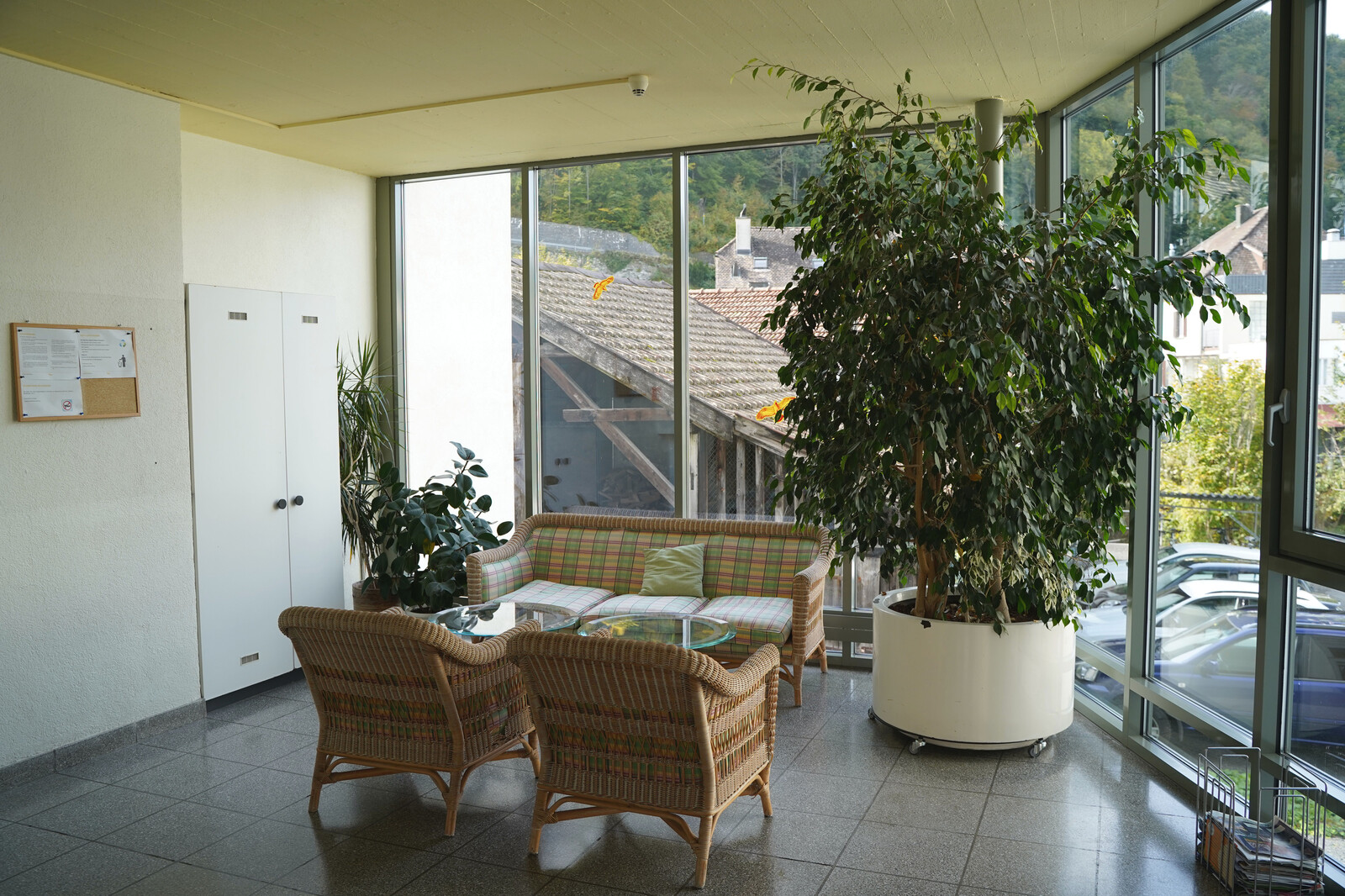 The common room of the Mühlebad staff house can be seen. In the common room of the staff house there are plants, armchairs and a sofa. On the opposite side of the street is the Hotel Grand Resort Bad Ragaz.
