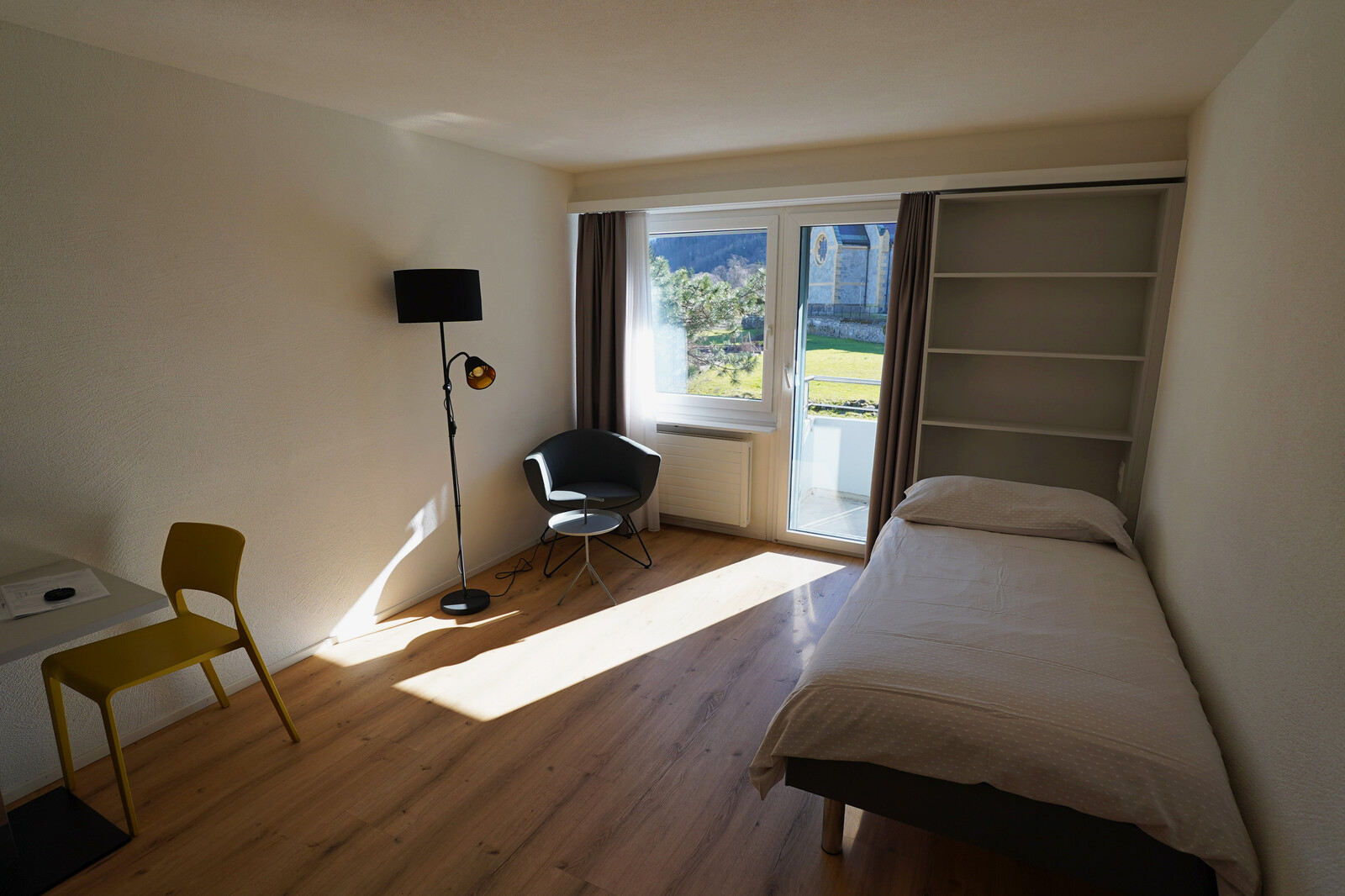 Mit einem Bett, einem Regal, einem Sessel, Tisch mit Stuhl und einem Blick nach draussen ist das Studio im Personalhaus Blumenrain ausgestattet. Es gehört zum Hotel Grand Resort Bad Ragaz und liegt in dem Dorf Bad Ragaz.