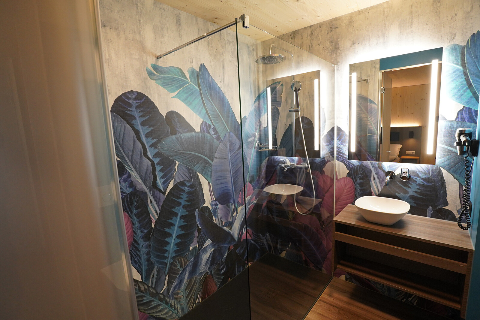Modern bathroom with large rainforest-themed wallpaper featuring vibrant blue and purple leaves. Glass shower, wooden vanity, and soft lighting.